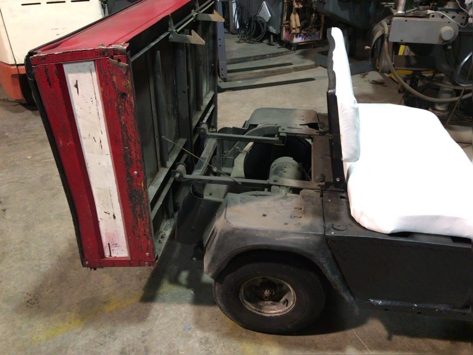 The EZGO's red dump bed tilted up, exposing the frame and drivetrain underneath