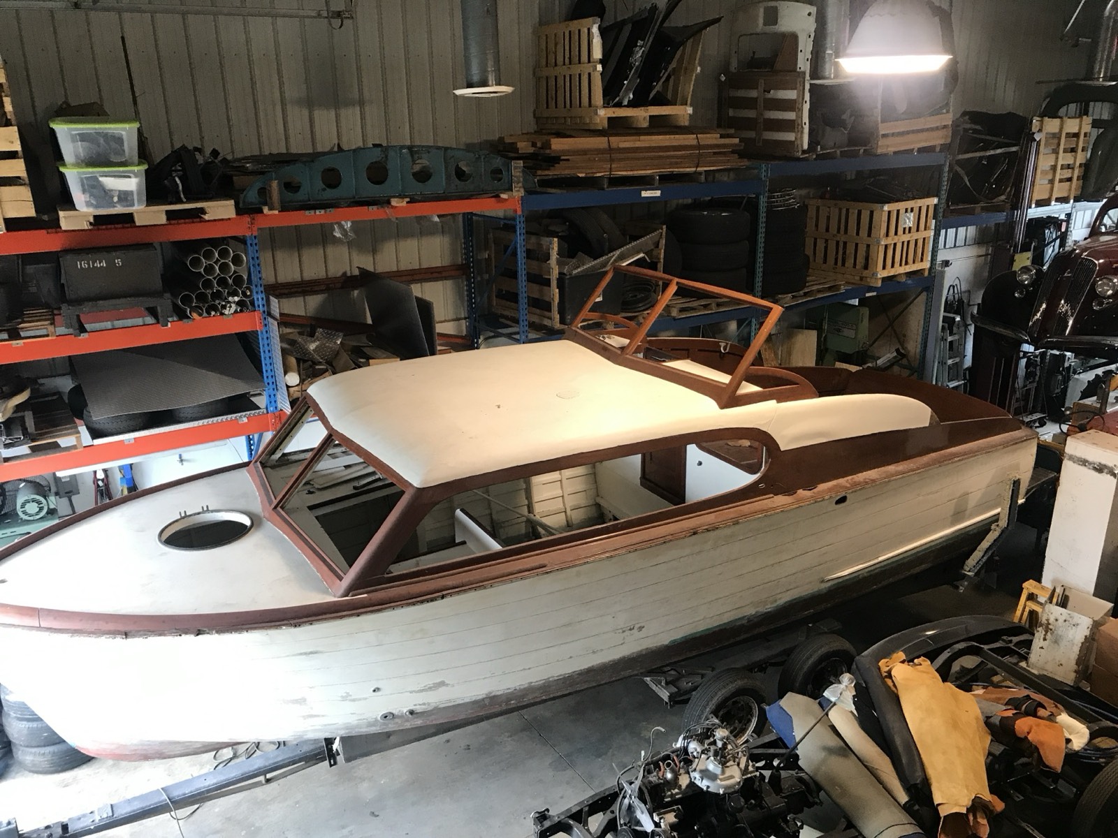 A Chris-Craft cabin boat inside Eric's shop with pallet racking loaded with parts and material behind it